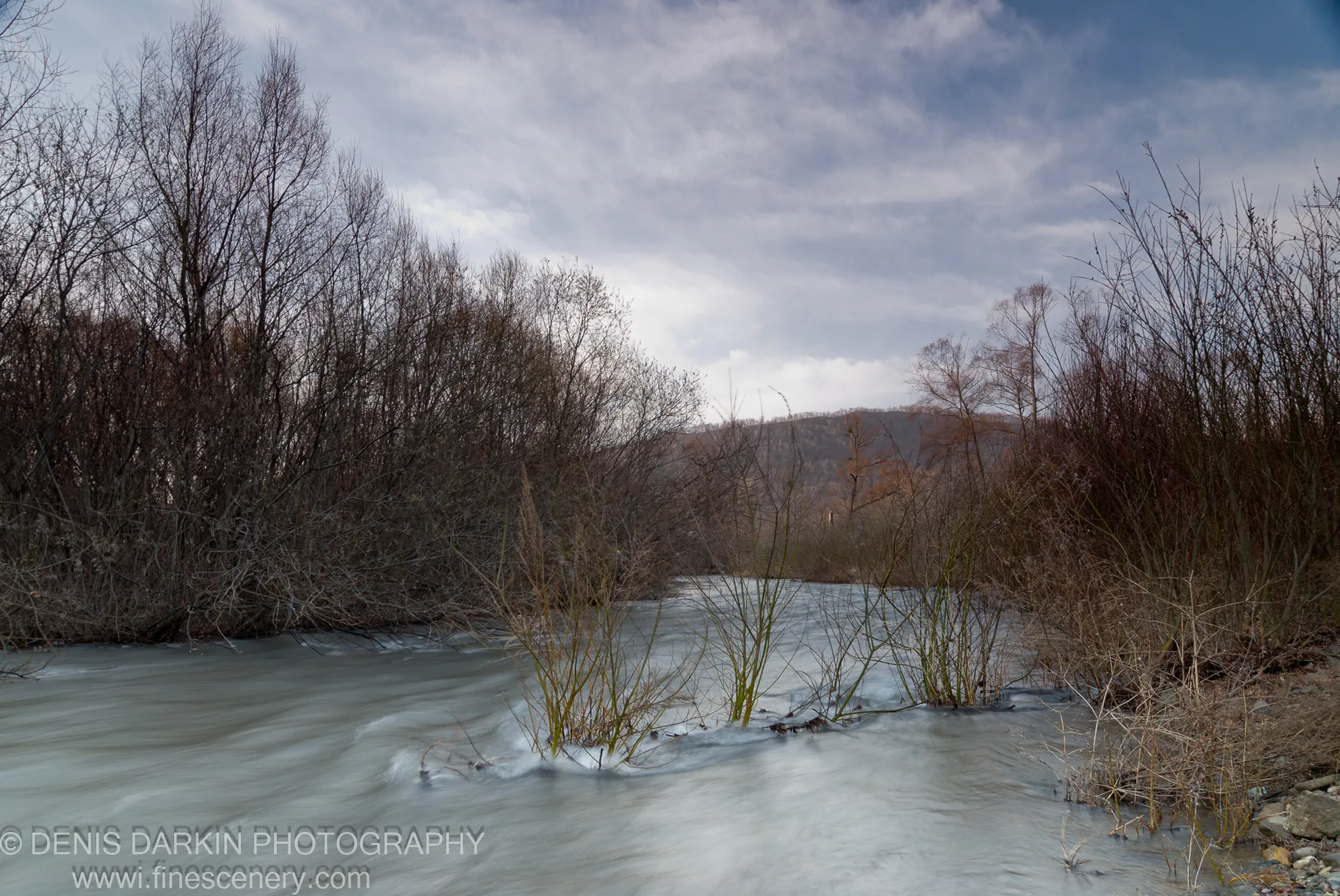 The Steklyanukha River in April, shot at slow shutter speed, with bare-branched trees lining both banks under an overcast sky.