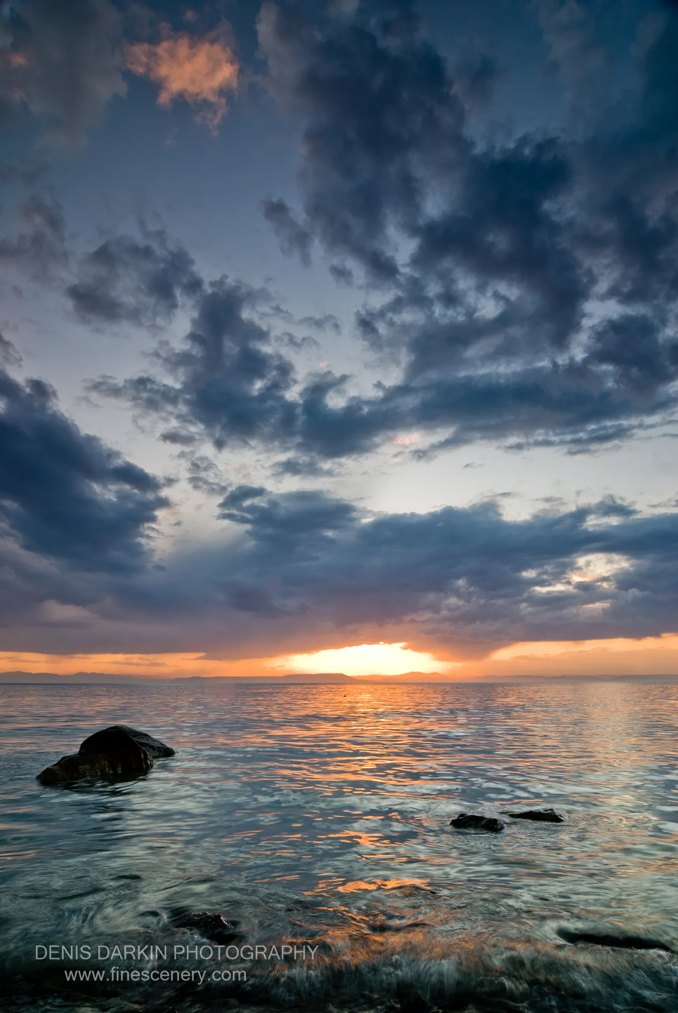 Dark coastal rocks sit in the foreground beneath a sky of storm clouds, warm light crossing the water at dusk, Vladivostok.