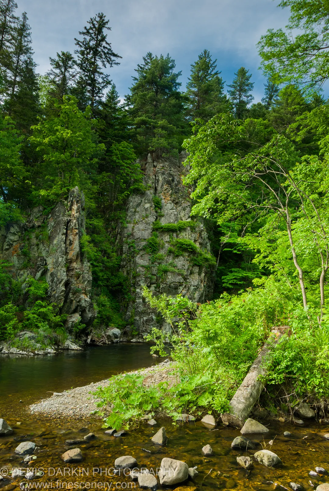 A clear shallow river flows alongside a vertical granite cliff face covered in moss and green foliage, Primorsky Krai, Russia.