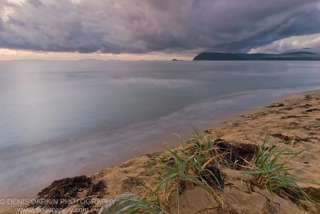 Tufts of beach grass and dark seaweed on a sandy shore at Sidemi Bay, with heavy clouds massing above the water in early morning.