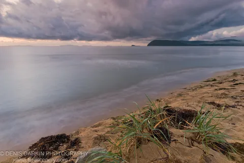 Tufts of beach grass and dark seaweed on a sandy shore at Sidemi Bay, with heavy clouds massing above the water in early morning.