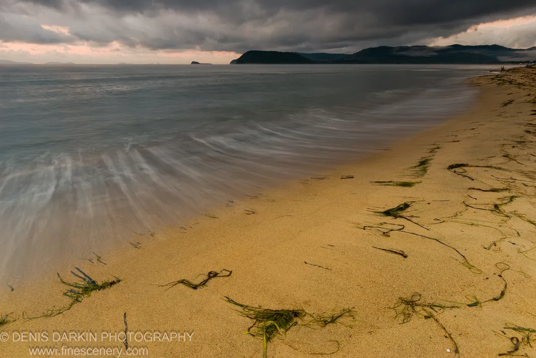 Motion-blurred morning waves wash over a sandy shore at Sidemi Bay, seaweed strands on the beach below a heavy bank of storm clouds.