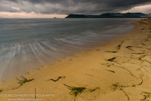Motion-blurred morning waves wash over a sandy shore at Sidemi Bay, seaweed strands on the beach below a heavy bank of storm clouds.
