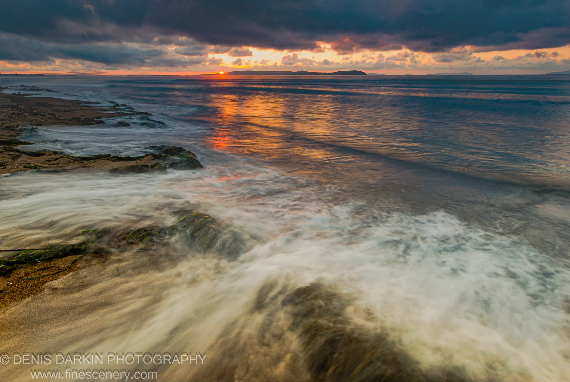 Long-exposure waves wash over flat dark rocks at Sidemi Bay, warm light breaking through heavy clouds in early morning.