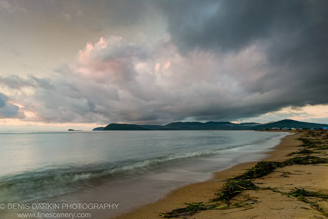 Smooth long-exposure waves wash over a seaweed-strewn sandy beach under heavy storm clouds with pink-tinted undersides at morning, Sidemi, Primorsky Krai.