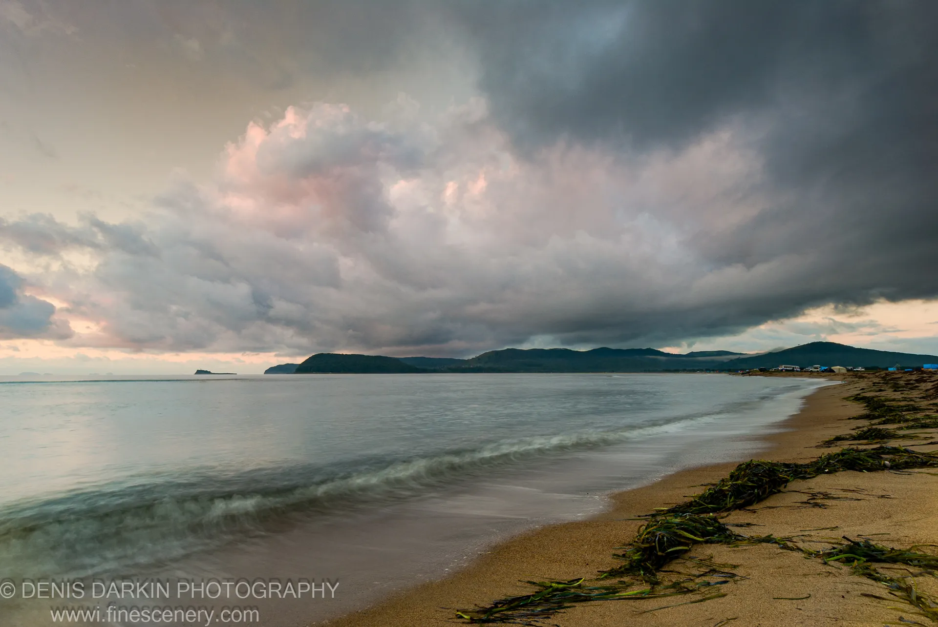 Smooth long-exposure waves wash over a seaweed-strewn sandy beach under heavy storm clouds with pink-tinted undersides at morning, Sidemi, Primorsky Krai.
