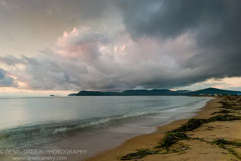 Smooth long-exposure waves wash over a seaweed-strewn sandy beach under heavy storm clouds with pink-tinted undersides at morning, Sidemi, Primorsky Krai.