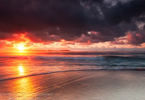 Intense morning light breaks through heavy dark clouds over Sidemi Bay, casting a golden path of reflection across the surf and wet sand below.