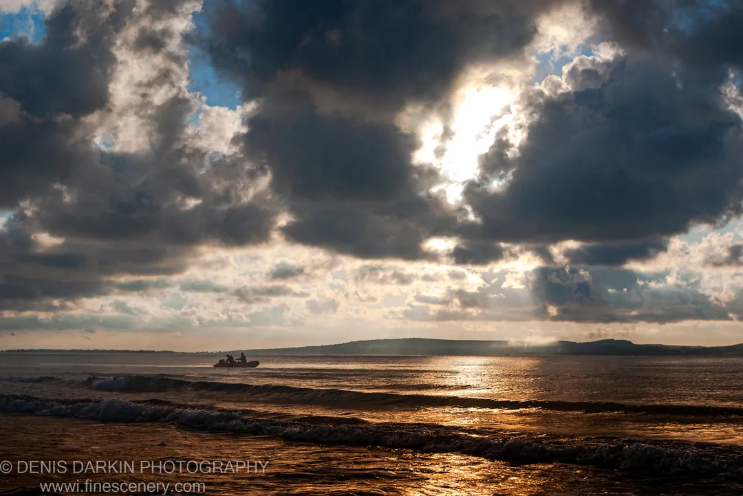 A small boat on Sidemi Bay in early morning, silhouetted against sunbeams breaking through heavy storm clouds above rolling waves.
