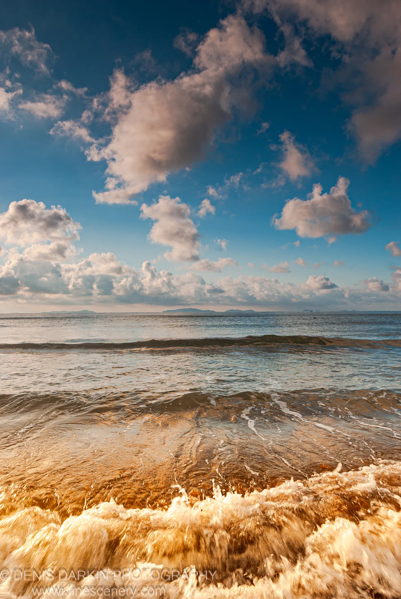 Breaking waves with heavy white foam roll onto the shore at morning against a deep blue sky with cumulus clouds, Sidemi coast.
