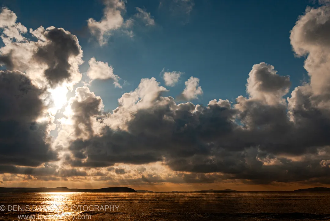 Backlit cumulus clouds with a sunburst at the horizon cast light onto dark water, with silhouetted headlands in the distance, Primorsky Krai coast.