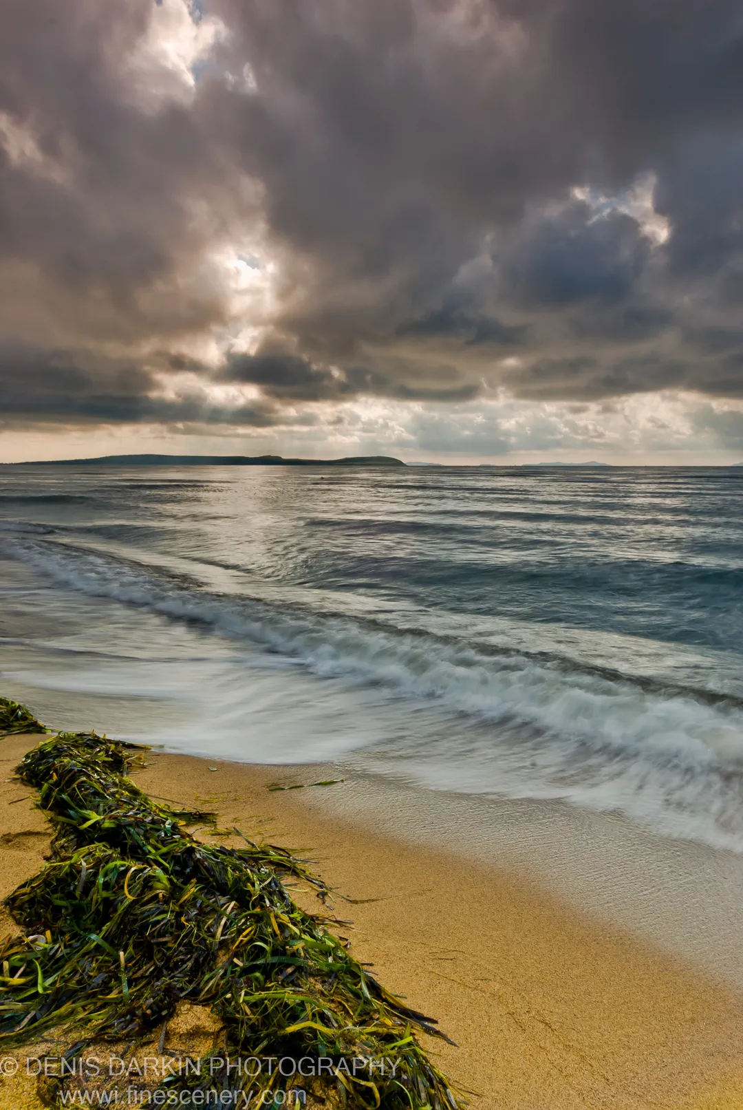 A mound of green kelp on wet sand at Sidemi Bay, long-exposure waves behind it and sunbeams through dark clouds above.