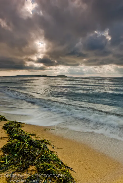 A mound of green kelp on wet sand at Sidemi Bay, long-exposure waves behind it and sunbeams through dark clouds above.