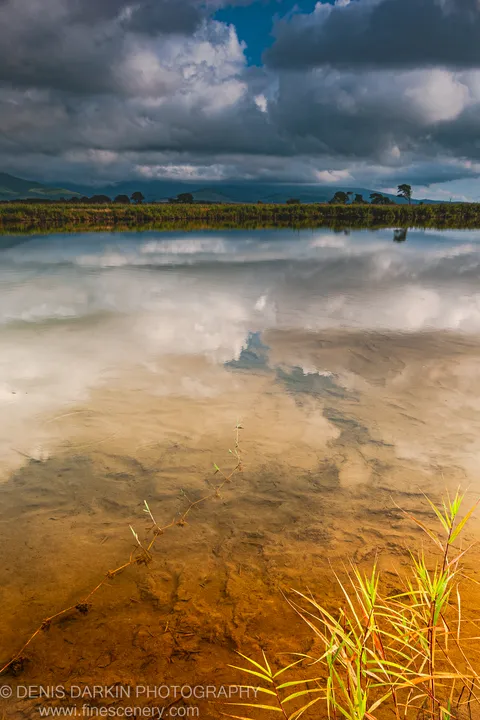 Delicate grasses grow from the sandy bottom of a calm shallow bay at Sidemi, their reflection in the still water below heavy cumulus clouds.