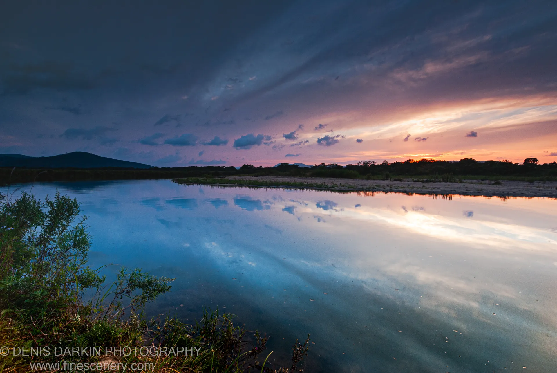 A calm lagoon at Sidemi mirrors a dusk sky with orange light on the horizon and dark silhouetted hills beyond.