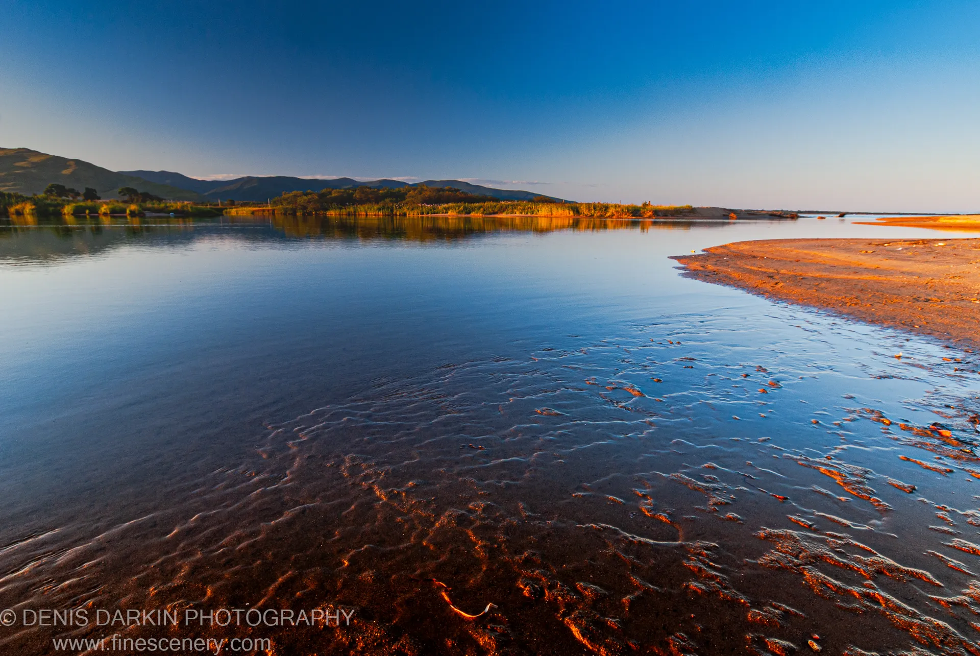 Wet sand ripples catch warm light at sunset on a curved sandy spit, with a reed-lined shore and distant hills beyond, Sidemi coast.