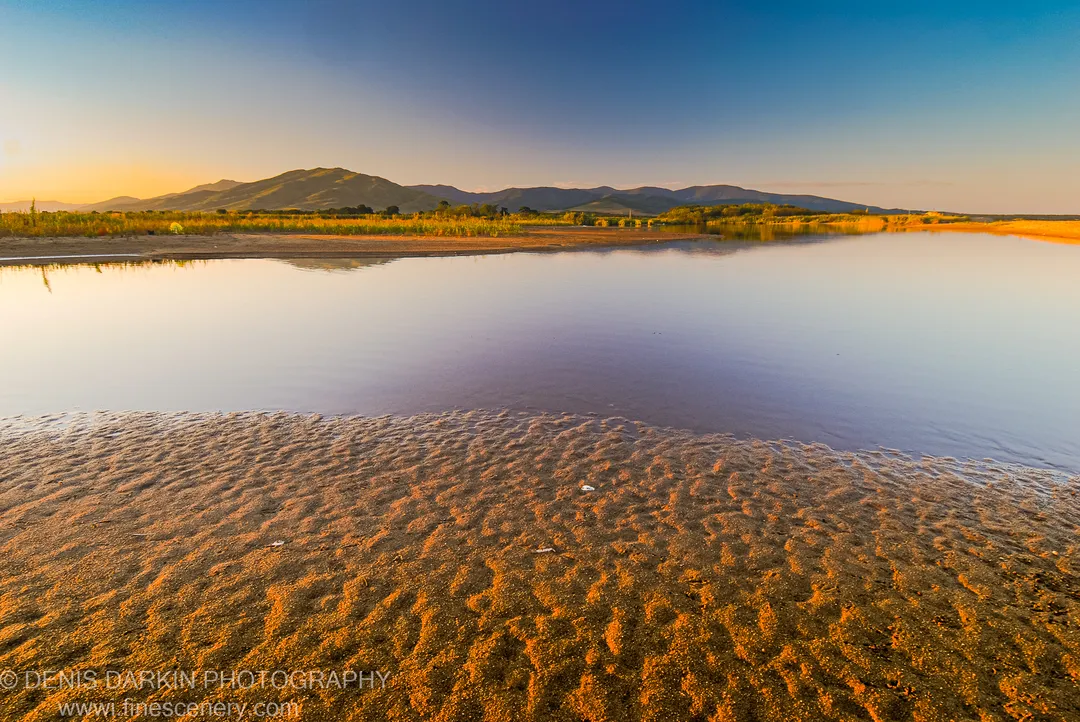 Sand ripples in evening light cross calm, glass-like water toward rolling hills under a gradient sunset sky, Hasan coast.
