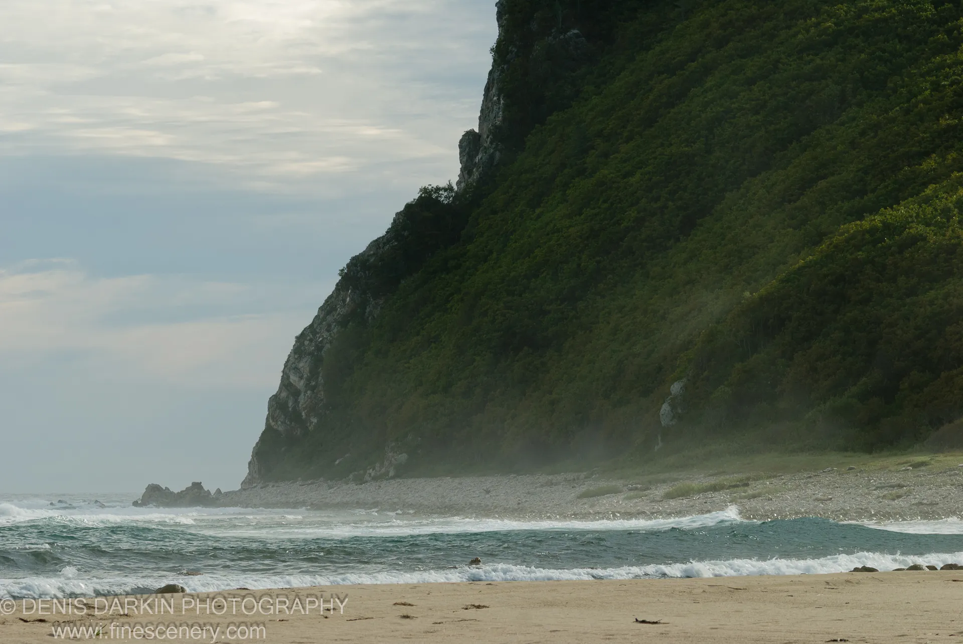 A densely forested cliff slopes toward a pebble shore where sea foam washes in, Lazovsky Reserve.