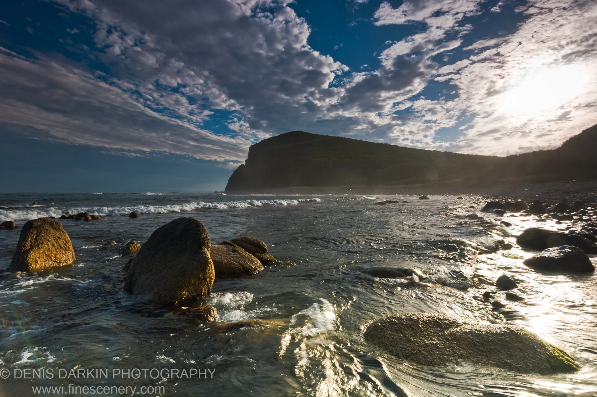 Wet boulders line the shore at Petrova Island as surf catches warm light, a steep forested headland silhouetted against textured clouds.