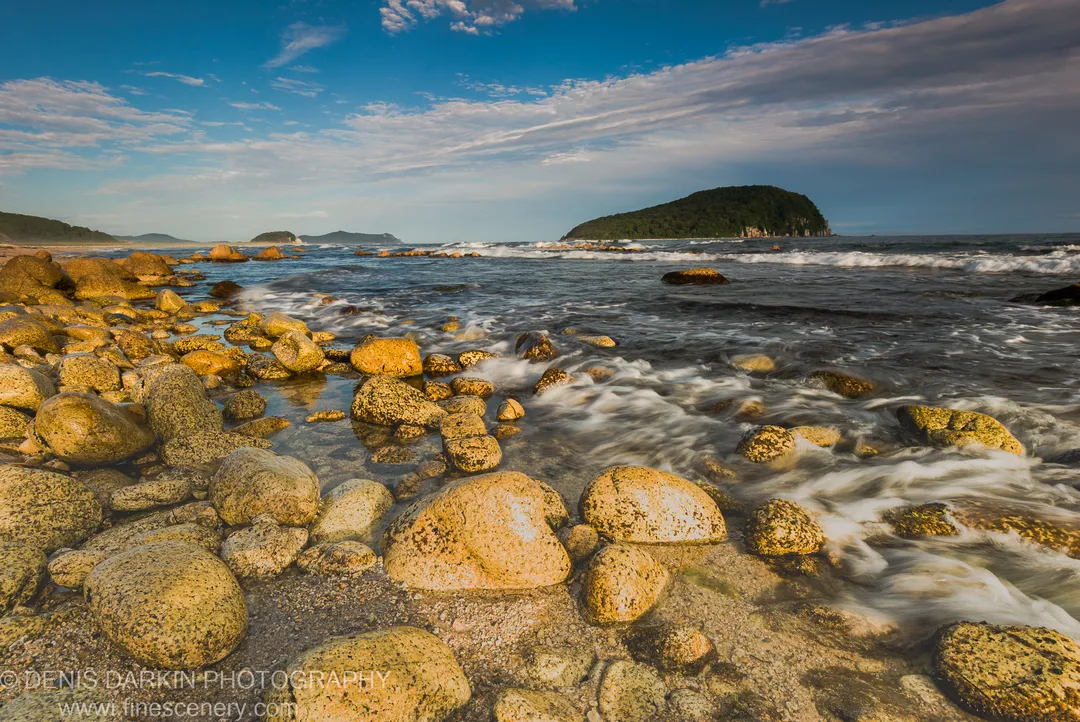 Smooth lichen-covered boulders line the rocky shore at Petrova Island, frothy waves lapping against them, a forested headland under wispy clouds.
