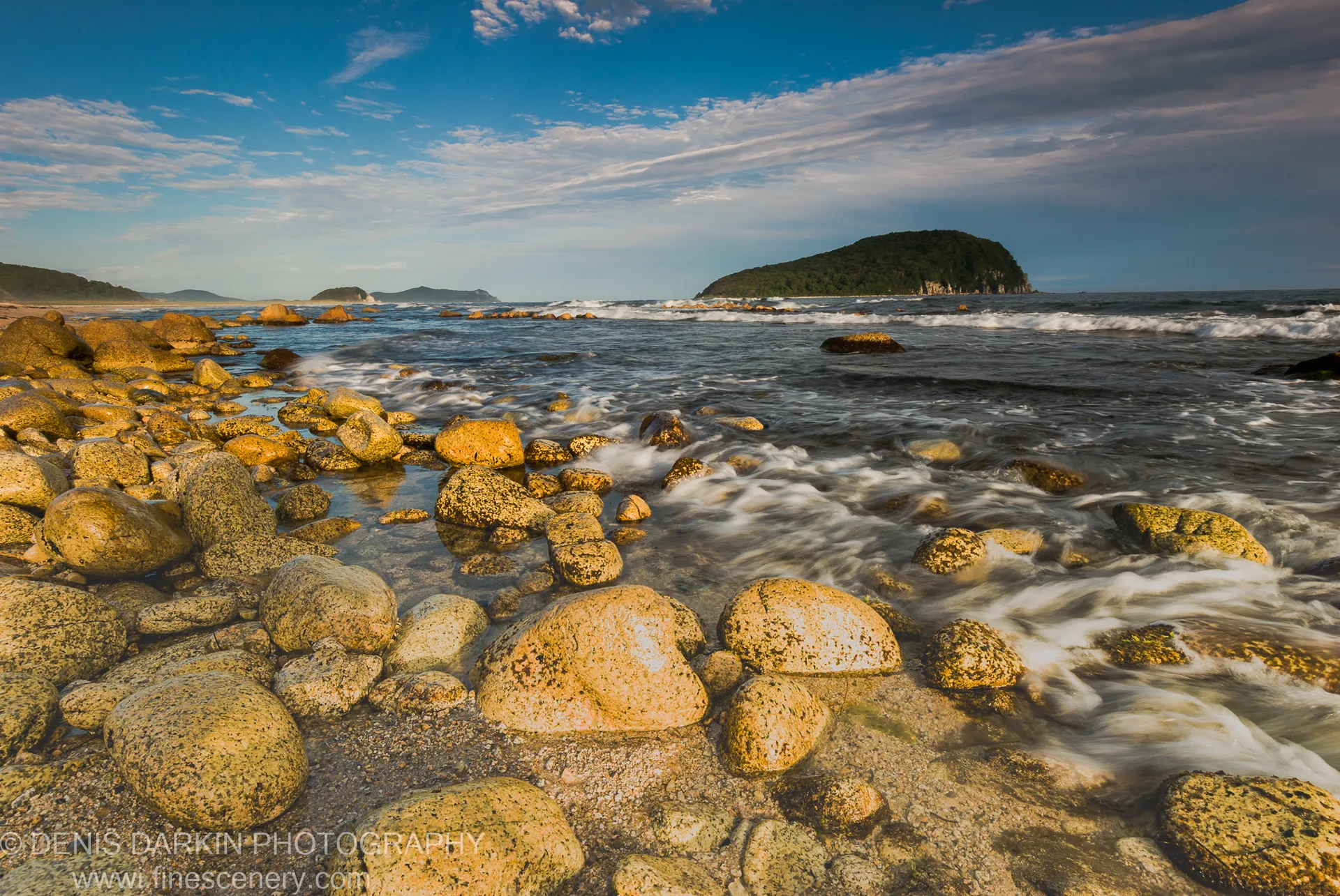 Smooth lichen-covered boulders line the rocky shore at Petrova Island, frothy waves lapping against them, a forested headland under wispy clouds.