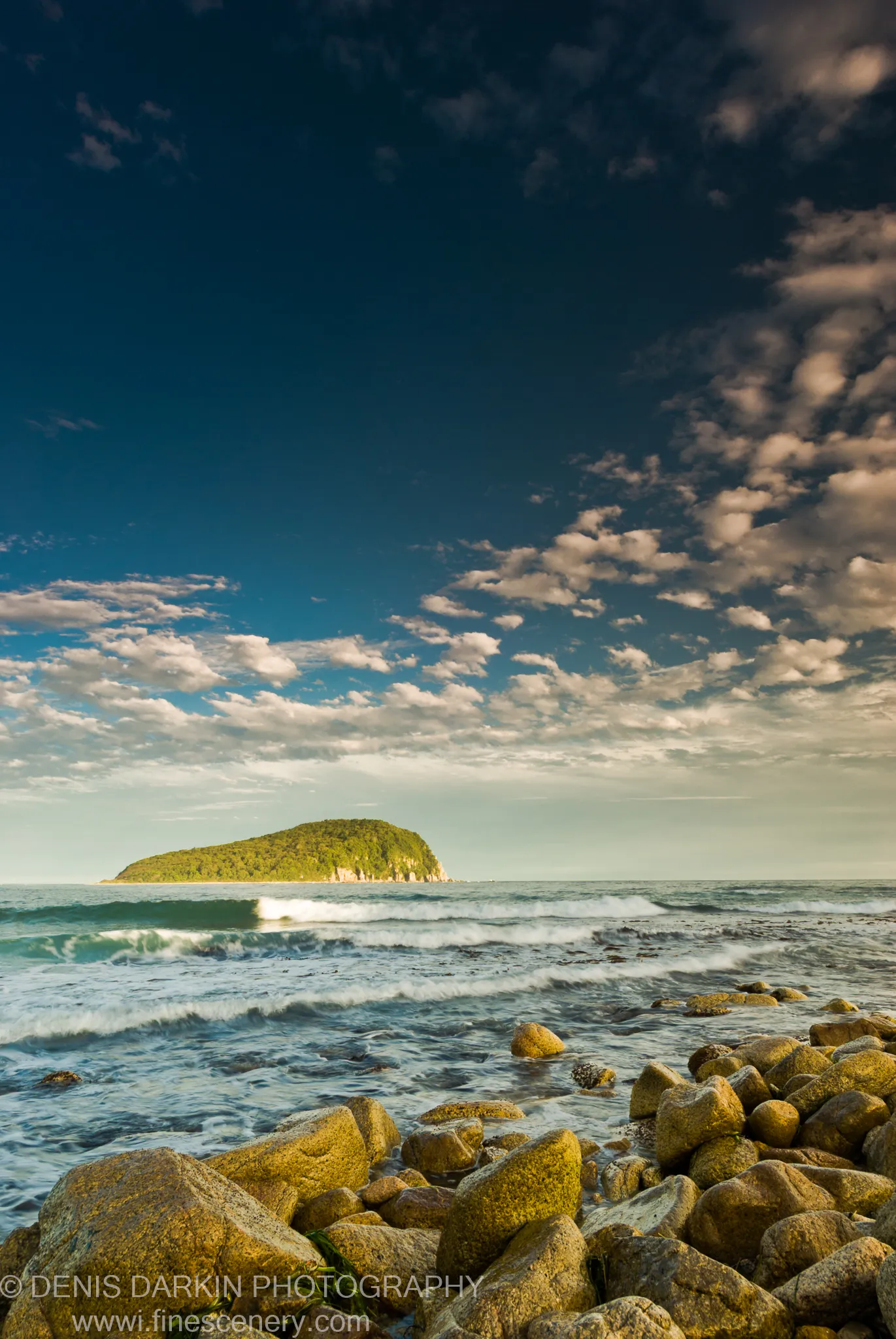 Moss-covered boulders in the foreground with a solitary forested island on the horizon, breaking waves between them, Lazovsky Reserve.