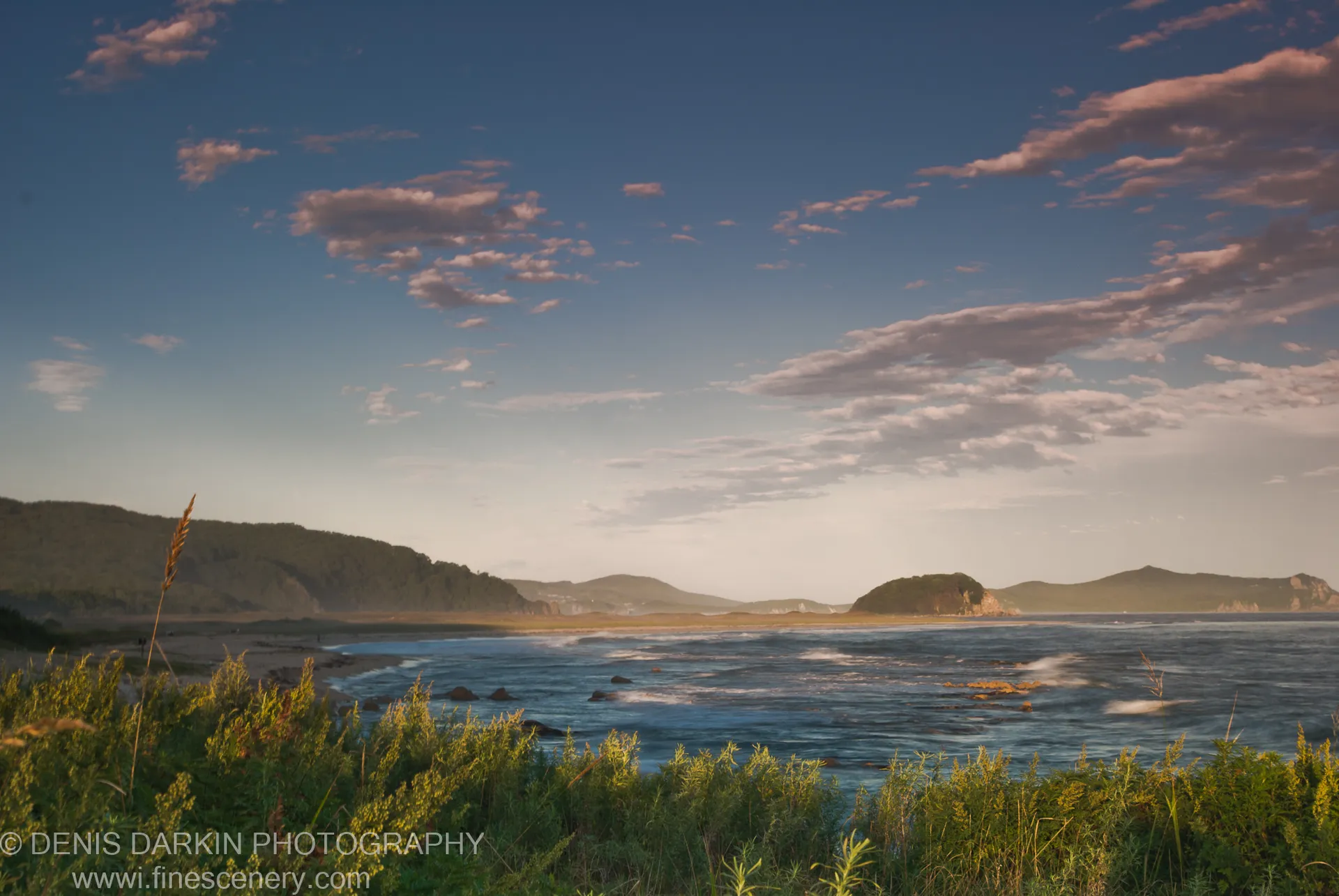 A coastal bay viewed across tall grass, scattered clouds above deep blue water and tree-covered hills, Lazovsky Reserve.
