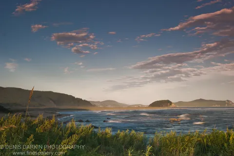 A coastal bay viewed across tall grass, scattered clouds above deep blue water and tree-covered hills, Lazovsky Reserve.