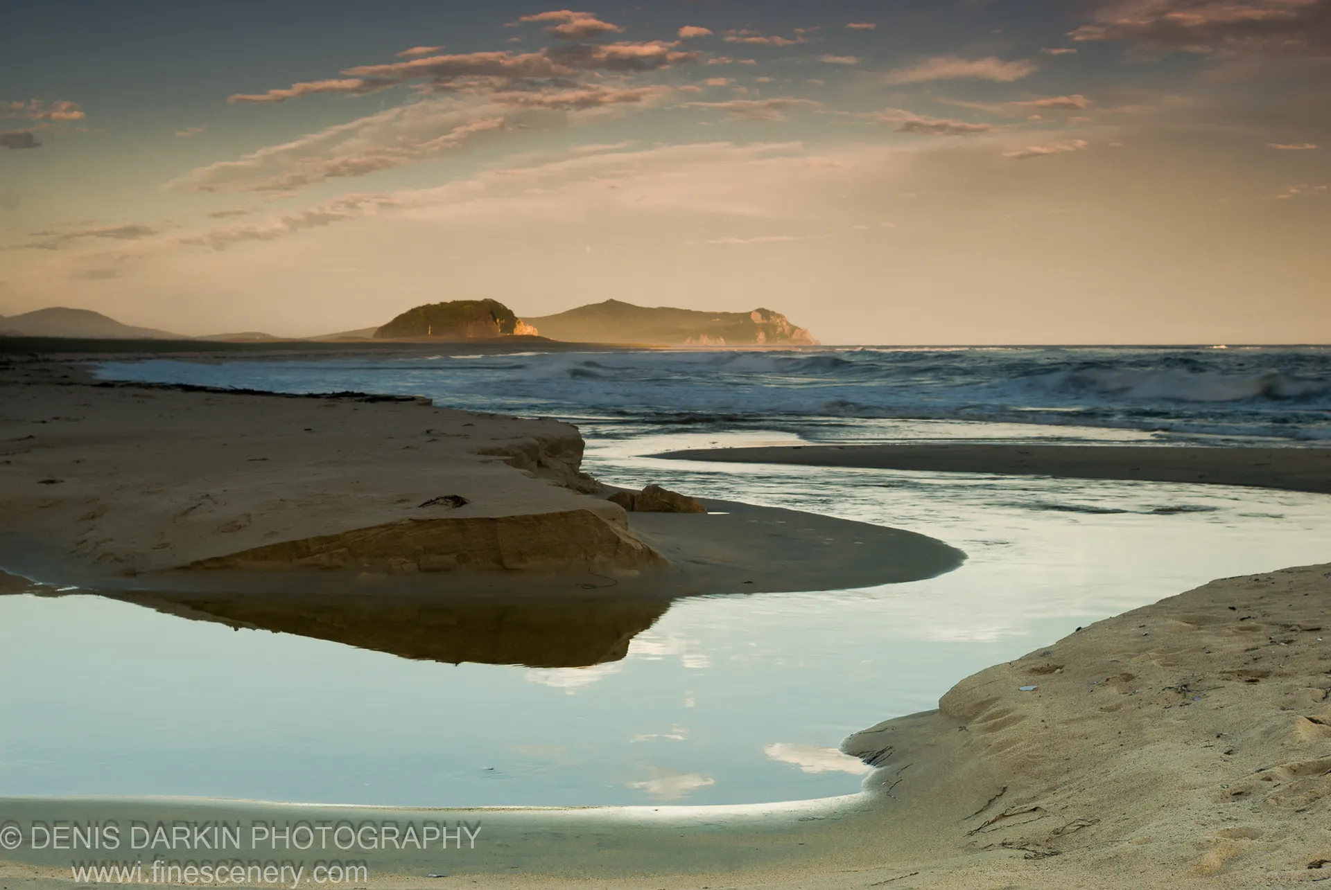 A tidal pool mirrors the sky and distant islands on a sand spit at Petrova Island, waves breaking beyond in warm golden light.