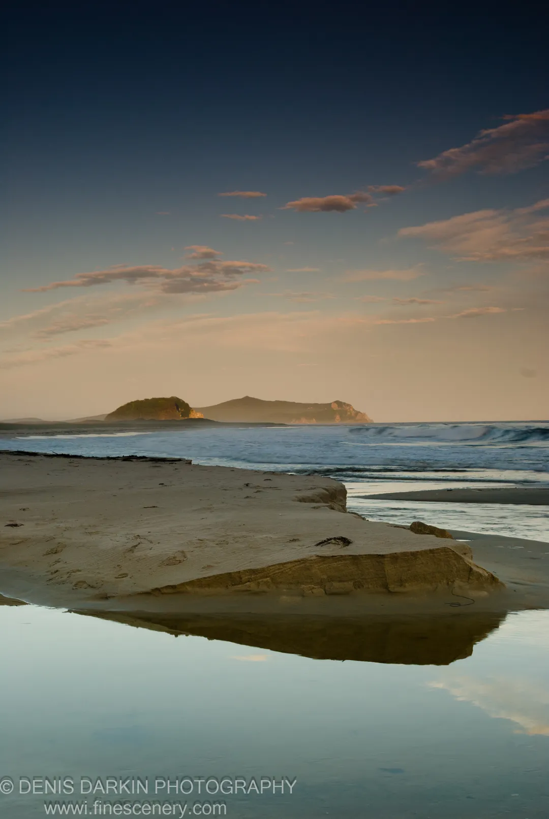 Sandy spit and tidal pool at Petrova Island, waves rolling toward shore under a sky fading from blue to soft peach near the horizon.