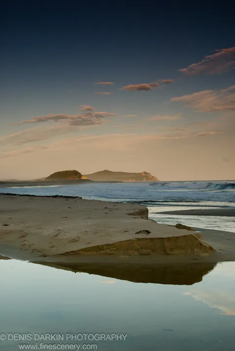 Sandy spit and tidal pool at Petrova Island, waves rolling toward shore under a sky fading from blue to soft peach near the horizon.