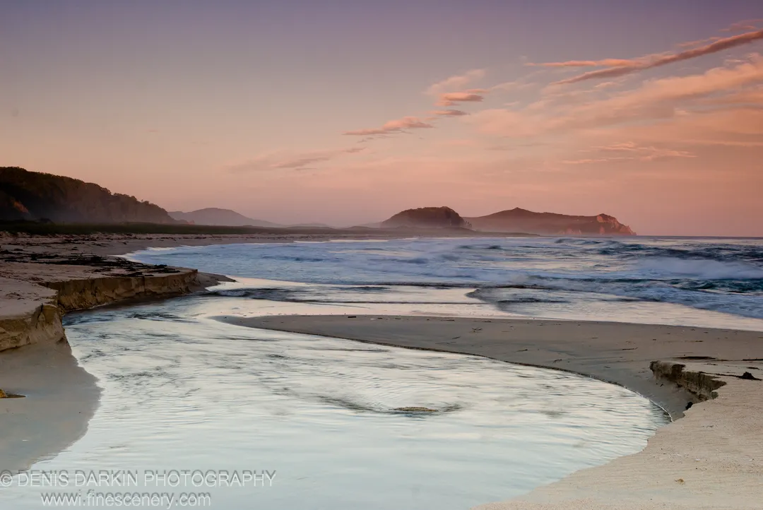 A stream meanders across a sandy beach into the ocean, reflecting pink and lavender clouds above silhouetted headlands, Lazovsky Reserve.