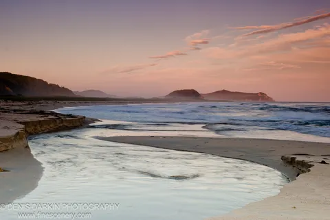 A stream meanders across a sandy beach into the ocean, reflecting pink and lavender clouds above silhouetted headlands, Lazovsky Reserve.