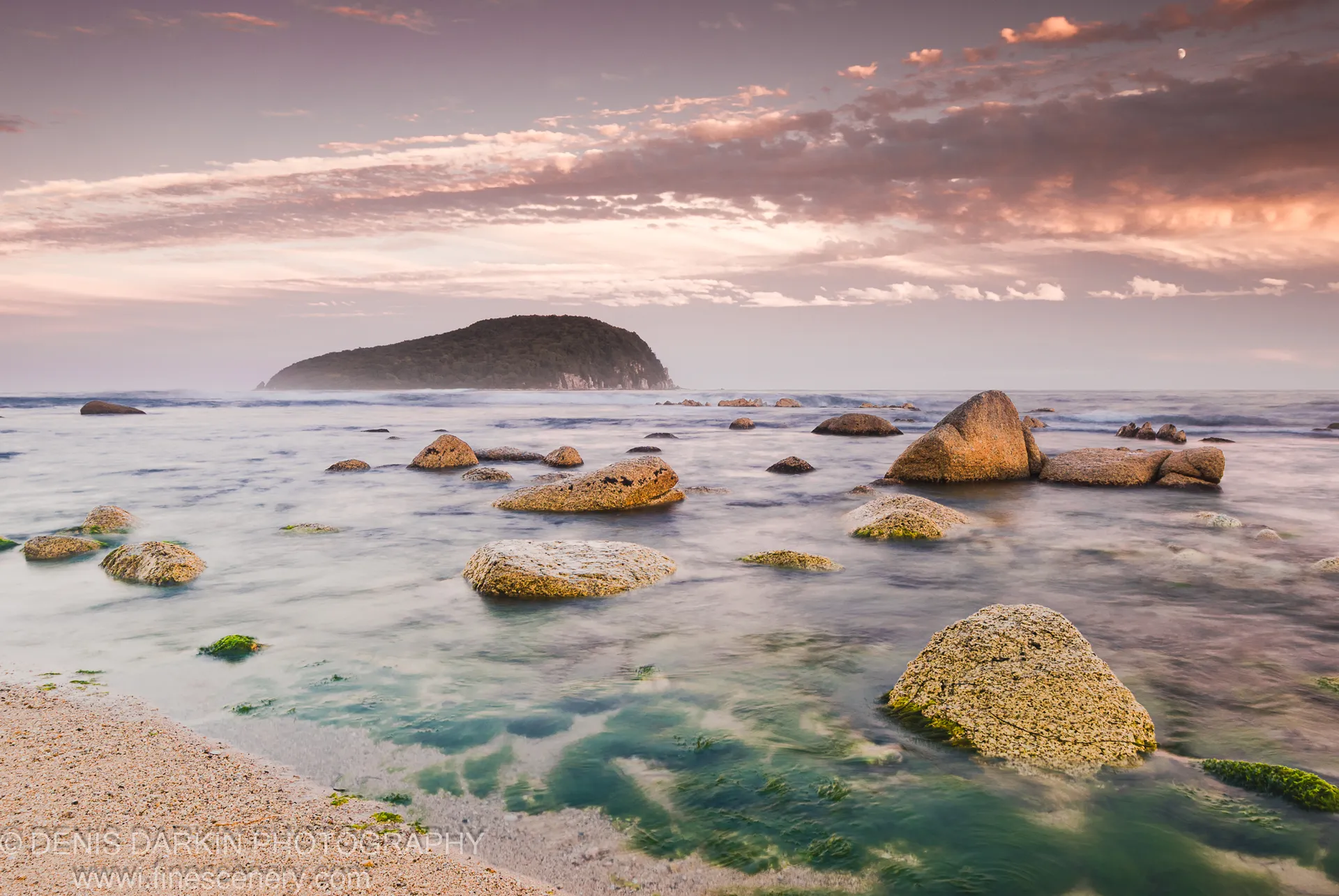 Moss-dusted boulders in still water at dusk at Petrova Island, a crescent moon above a pastel sky of pink and lavender over the forested headland.