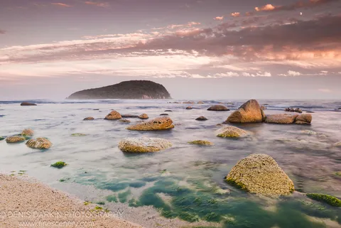 Moss-dusted boulders in still water at dusk at Petrova Island, a crescent moon above a pastel sky of pink and lavender over the forested headland.