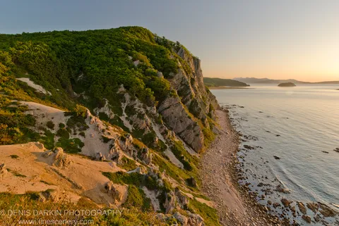 Morning sunlight on a rugged cliffside where dense forest meets exposed limestone and sandy slopes above calm coastal water, Lazovsky Reserve.