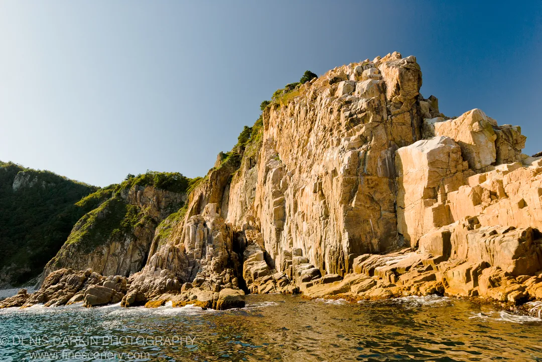 Sun-drenched coastal cliffs rise steeply from the ocean, their rock faces textured with moss and shadow, under a clear blue sky.