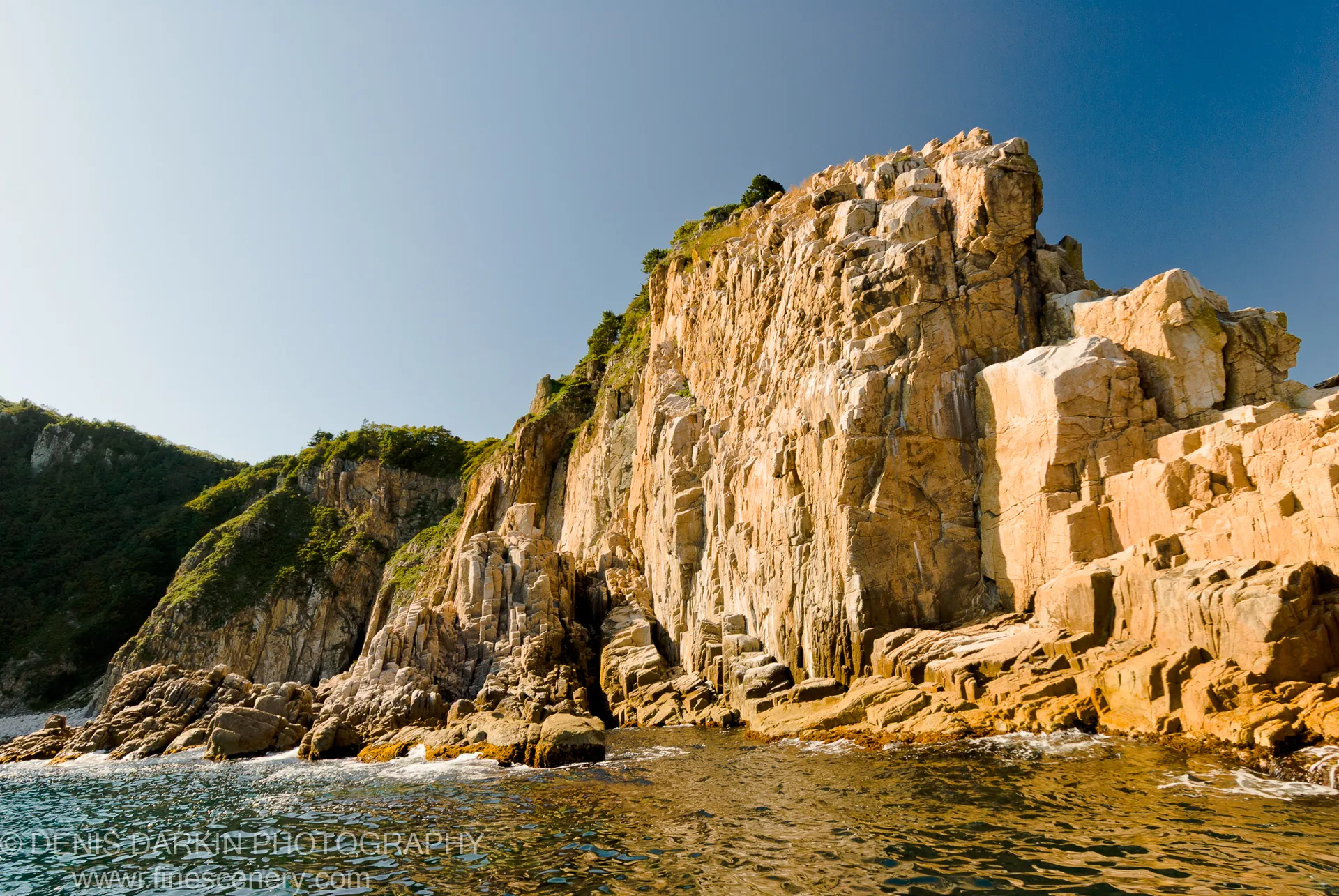 Sun-drenched coastal cliffs rise steeply from the ocean, their rock faces textured with moss and shadow, under a clear blue sky.