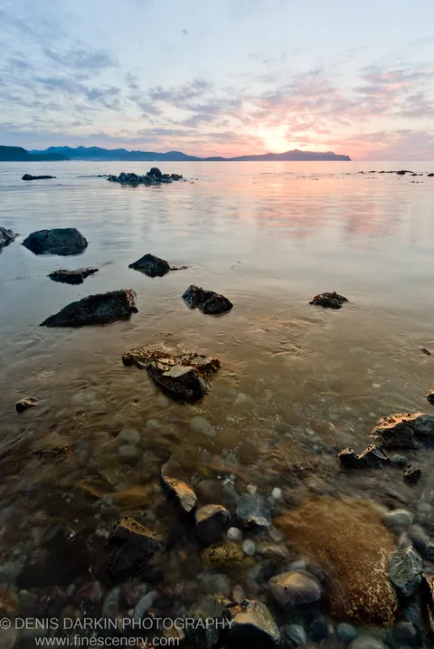 Smooth stones and mossy boulders beneath shallow transparent water at Preobrazheniye, silhouetted coastal hills under a pastel morning sky.
