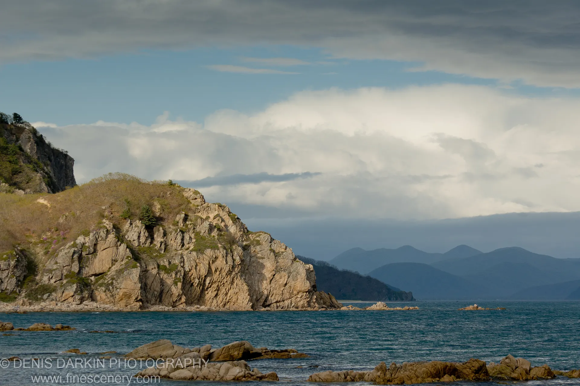 A tan stone headland under large cumulus clouds at golden hour, with hazy blue mountains visible across the water, Petrova Island.