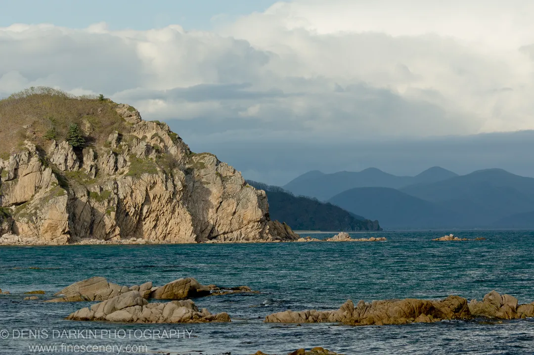 Steep tan cliff face rising from the sea at Petrova Island, with scattered rocky outcrops and distant mountains fading into cloud.