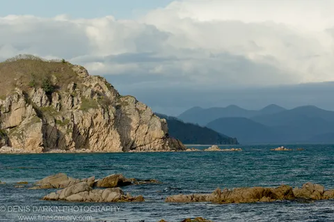 Steep tan cliff face rising from the sea at Petrova Island, with scattered rocky outcrops and distant mountains fading into cloud.