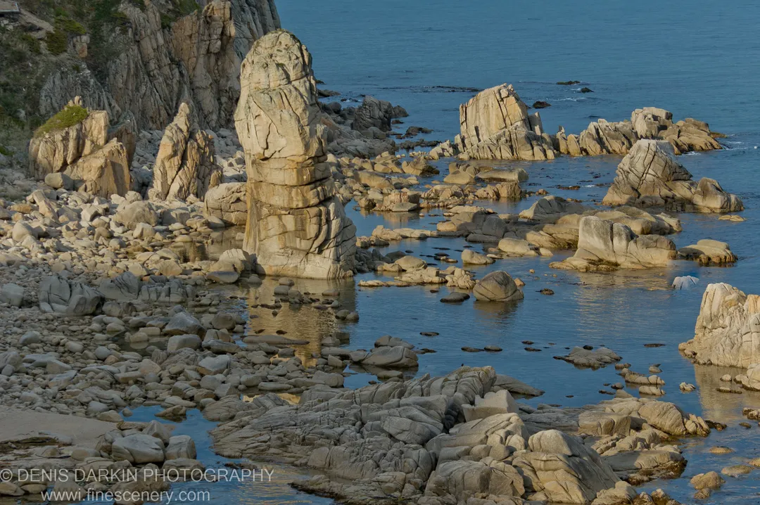 A tall sea stack lit in warm light at Petrova Island, with scattered boulders and shallow reflective pools in the foreground.