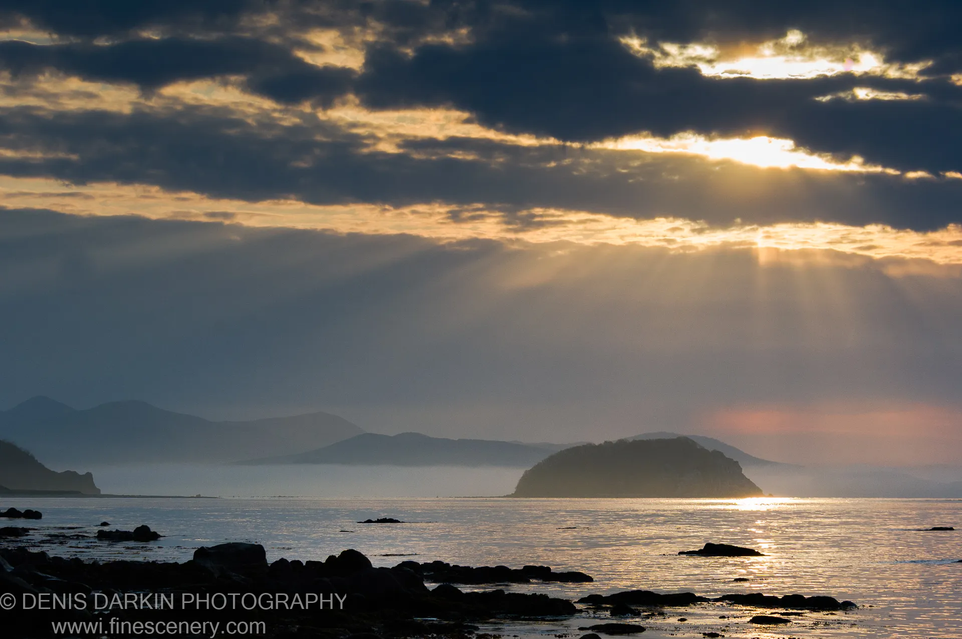 Golden morning rays breaking through dark clouds above a calm sea at Petrova Island, a silhouetted island visible in the hazy distance.