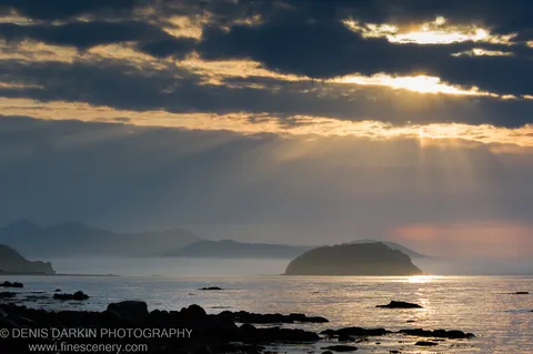 Golden morning rays breaking through dark clouds above a calm sea at Petrova Island, a silhouetted island visible in the hazy distance.