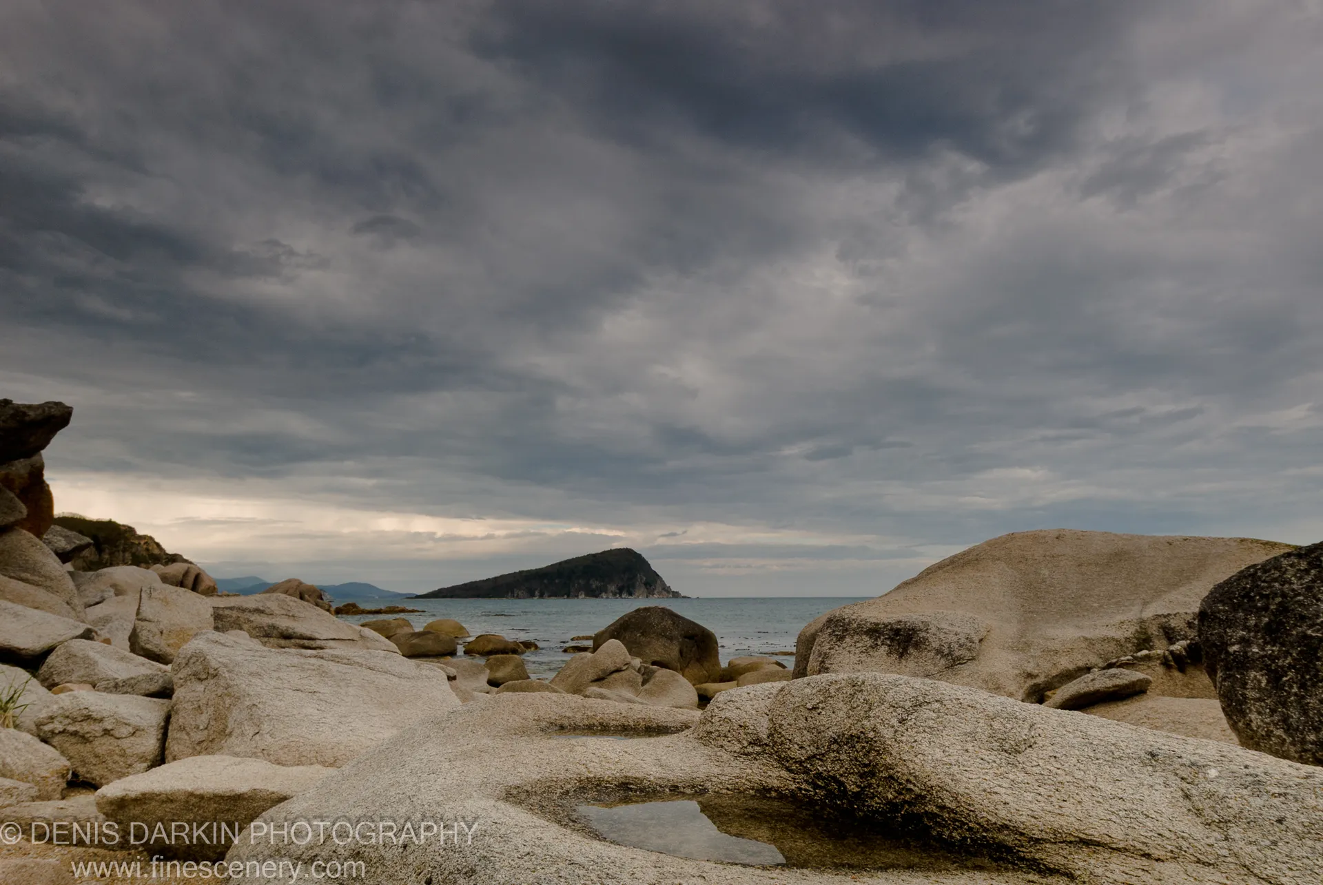 Textured granite boulders at Petrova Island with a dark headland and storm clouds overhead, diffuse light unifying stone and sea.