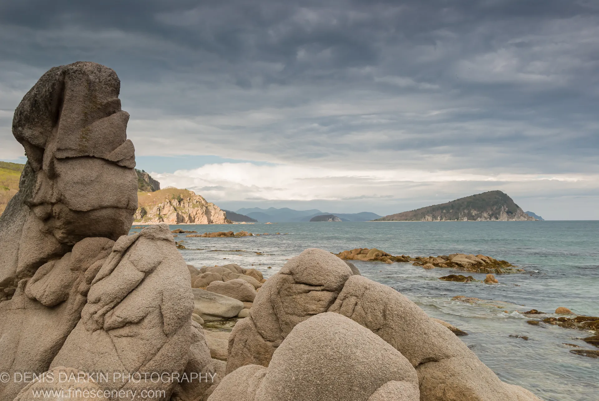 Coarse granite boulders on the rocky shore of Petrova Island with hazy offshore islands and mountain ranges emerging beneath grey cloud cover.