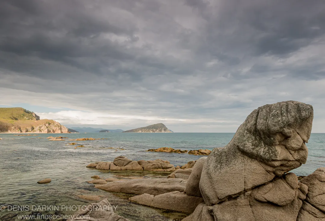 A large rock formation at the edge of a sheltered bay on Petrova Island, with dark seaweed underwater and a small island on the horizon.