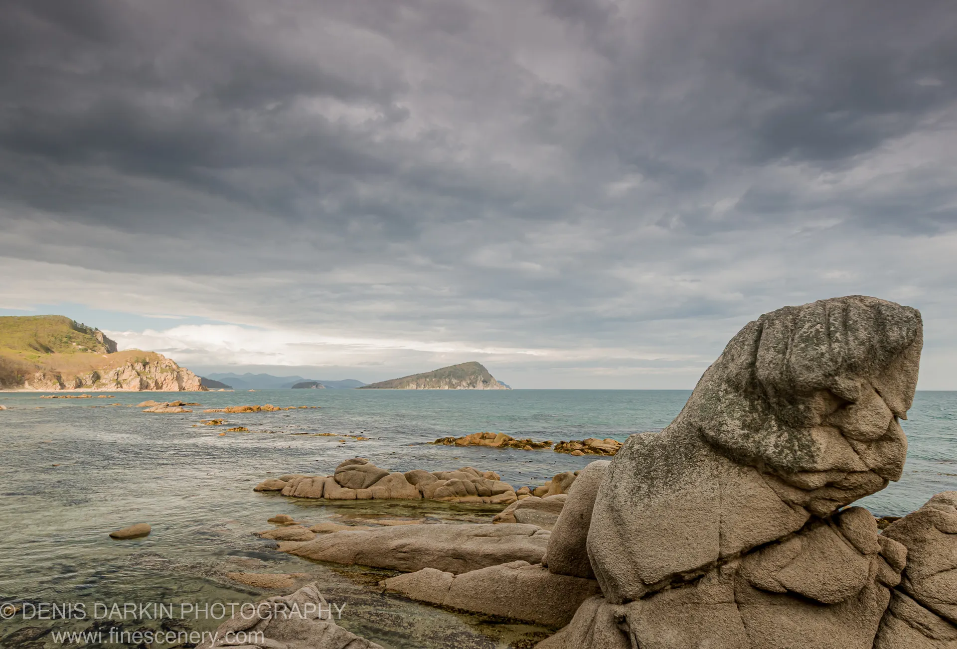 A large rock formation at the edge of a sheltered bay on Petrova Island, with dark seaweed underwater and a small island on the horizon.