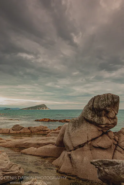 Large, moss-covered granite boulders on the coast of Petrova Island, with a solitary island visible across the calm sea beneath grey clouds.